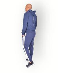 A man wearing navy blue and white side panel pants with zippers at legs and with a logo on the back of the leg, paired with a navy full zipped hooded jacket.