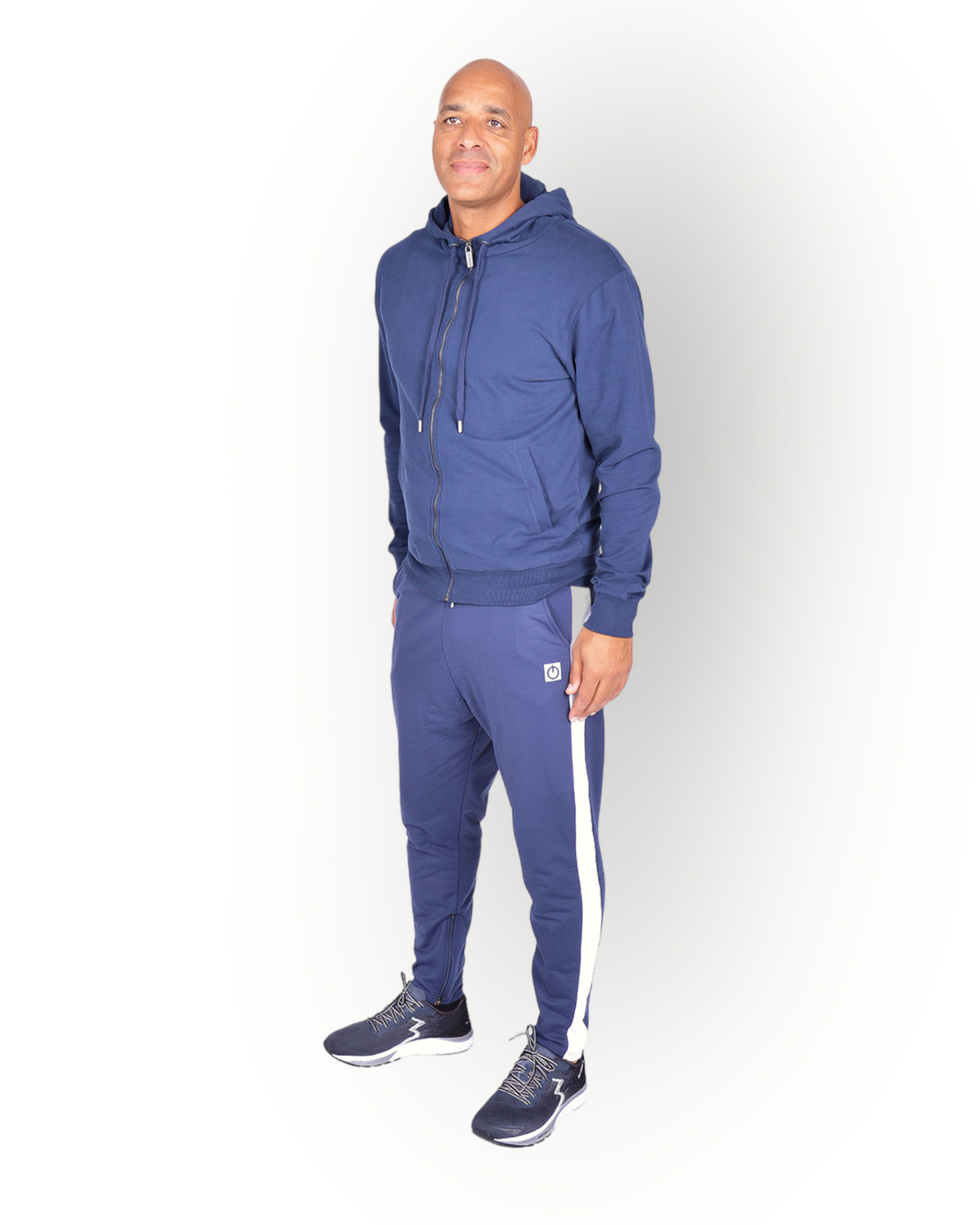 A man wearing navy blue and white side panel pants with zippers at legs and with a logo on the back of the leg, paired with a navy full zipped hooded jacket.