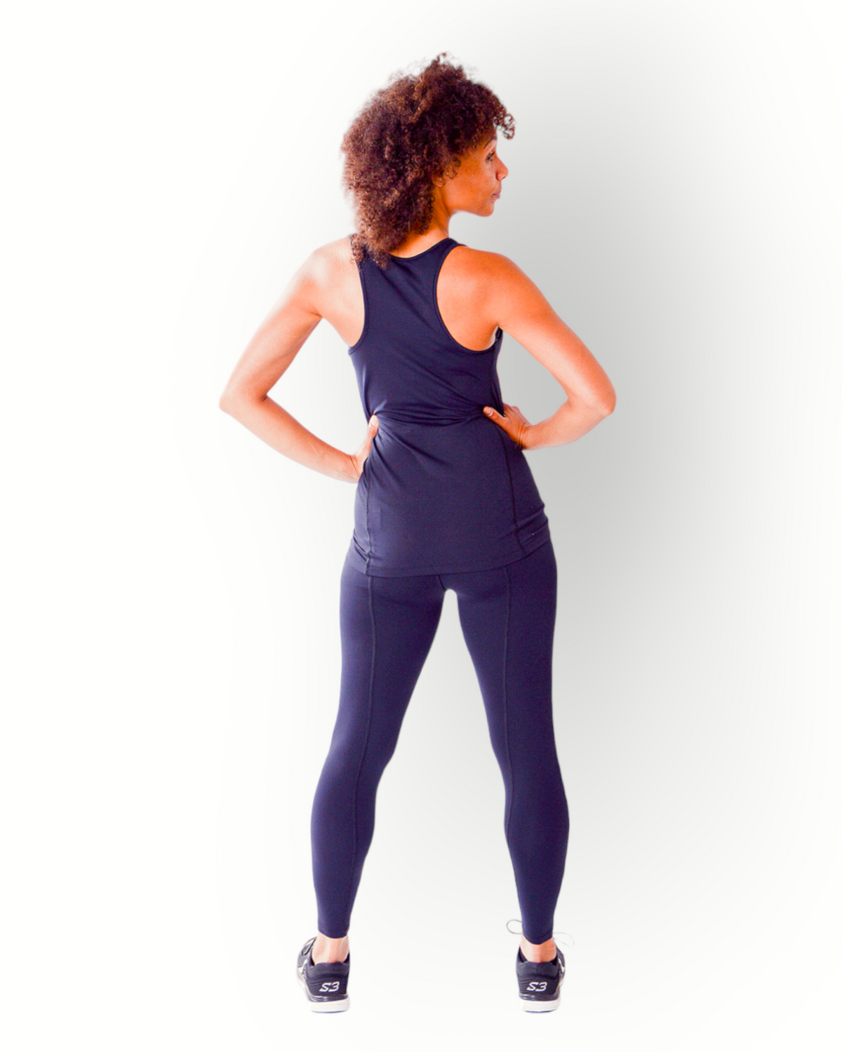 A woman wearing a black tank top with a racerback, flatlock seams and longer fit standing with hands on hips against a white background.