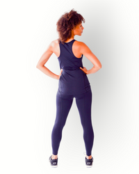 A woman wearing a black tank top with a racerback, flatlock seams and longer fit standing with hands on hips against a white background.