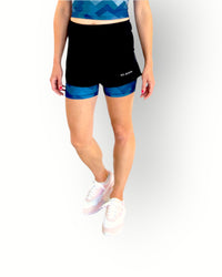 A woman wearing a black sports short with a camo blue print layer underneath. The short has an overlay fabric and an inner short, and there is a small pocket with zipper at the back inside the waistband.