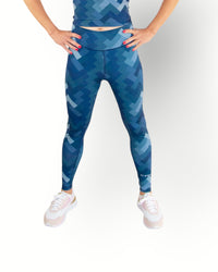 A woman standing, wearing reversible sport legging with  a geometric blue pattern showing and the other side is in solid navy color.